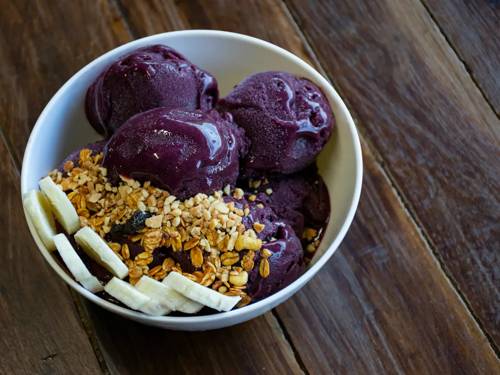 Weiße Schale mit Kugeln aus dunkellila Açaí, Granola und Bananenscheiben auf dunklem Holztisch