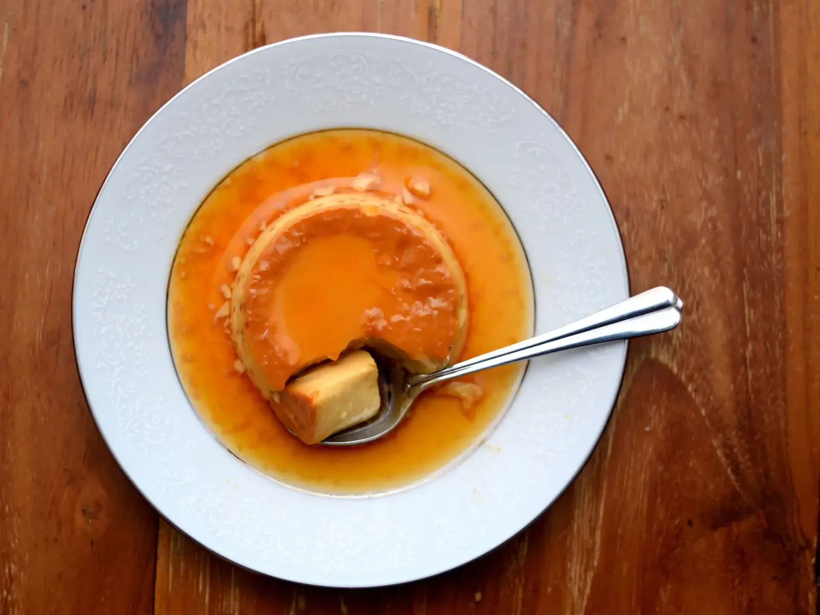 Karamellpudding mit Löffel auf einem weißen Teller auf Holztisch, von oben